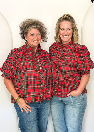 Two women standing side-by-side wearing matching red and black plaid puff-sleeve button-up shirts and blue jeans, posing in front of a white studio backdrop with arched panels.