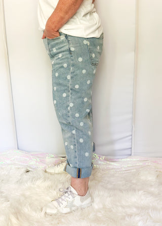 A person wearing light blue denim jeans with white polka dots, paired with a white t-shirt and white sneakers, standing on a white faux fur rug.