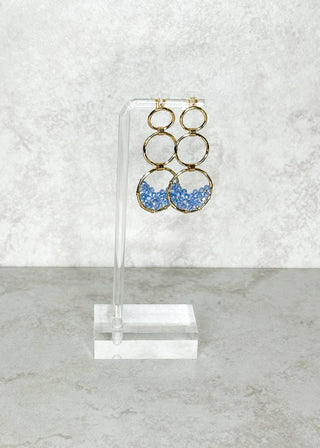 Gold and blue earrings on a clear stand against a light gray background