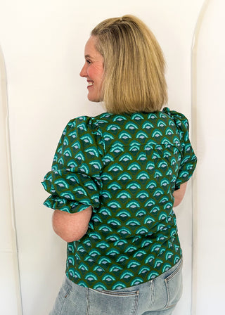 A woman with blonde hair is shown from the back wearing a dark green short-sleeve blouse with a repeating teal arch pattern, paired with light-wash jeans, standing against a white arched backdrop.