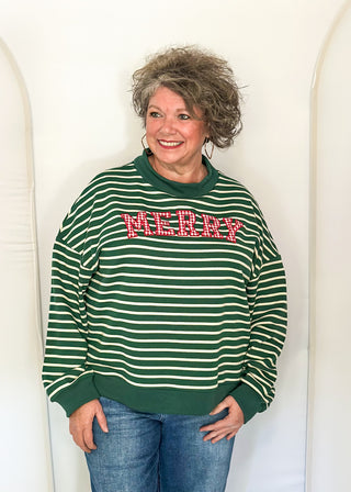 A woman with curly gray hair wearing a green and white striped crewneck sweatshirt with 'MCCRR' in red block letters, paired with blue jeans, standing against a white arched backdrop.
