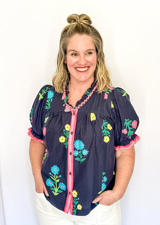 Navy floral short sleeve blouse with pink, green, yellow, and blue flowers, featuring puff sleeves, ruffled neckline, button front with pink trim, and a relaxed fit