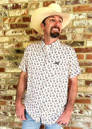A man wearing a cream cowboy hat and a white short-sleeve button-up shirt with a brown all-over floral print, standing in front of a rustic brick wall.
