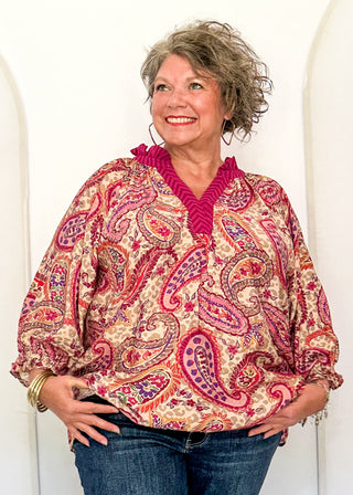 A smiling woman with short gray hair models a long-sleeve paisley blouse with a vibrant magenta V-neck trim, paired with blue jeans, standing in front of a white arched backdrop.