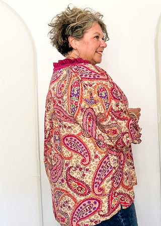 A woman with short gray hair is shown from the side, wearing a long-sleeve paisley blouse with a red, pink, and blue pattern on a beige background, paired with dark blue jeans. She is standing in front of a white arched backdrop.