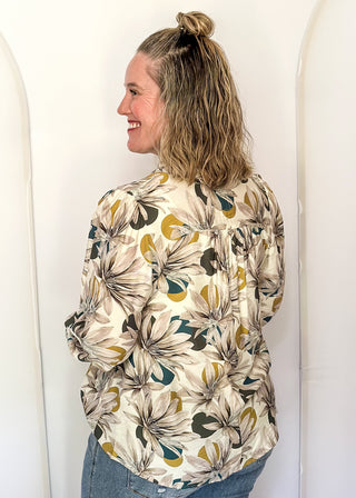 A woman wearing a cream-colored long-sleeve button-up shirt with a large floral print in olive green, mustard yellow, and black, paired with blue jeans, standing against a white arched backdrop.