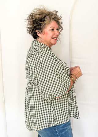 A woman wearing a green and white gingham button-up shirt and blue jeans, standing in a studio with a white arched backdrop.