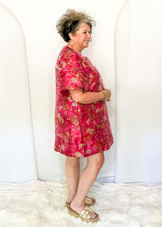 A woman modeling a short-sleeve pink dress with a gold floral pattern, standing in front of a white arched backdrop.