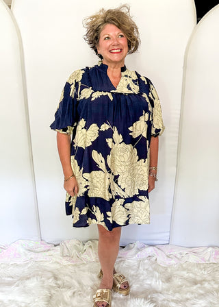 Woman wearing a navy and gold floral dress standing against white cylindrical objects.