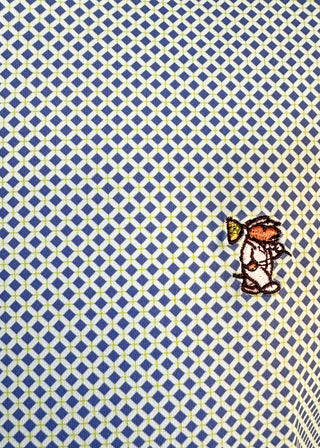A close-up of a patterned fabric featuring a repeating blue and white diamond lattice with thin yellow lines, accented by a small embroidered character motif in orange, brown, and yellow near the lower right.