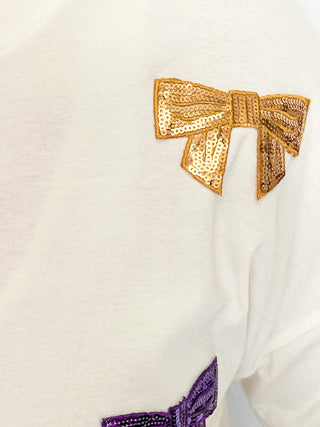 close up of white top featuring scattered sequin bows in gold and purple