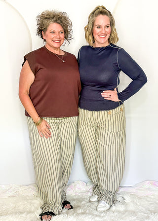 Two women standing side-by-side in a studio setting, wearing matching beige and white vertically striped pants. The woman on the left wears a brown sleeveless top, and the woman on the right wears a dark navy long-sleeve top.