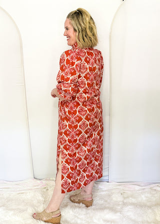 A woman wearing a red and white patterned long-sleeve midi dress, standing in profile on a white faux fur rug against a white studio backdrop with arched panels.