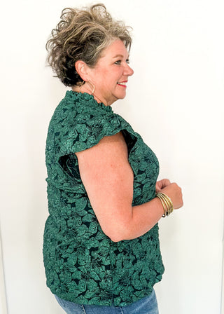 side view of woman wearing forest green jacquard flutter sleeve blouse with a floral pattern