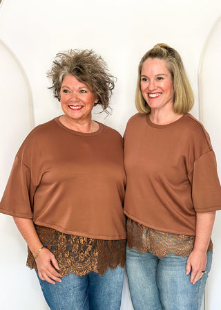 Two women standing side by side wearing matching brown short-sleeve t-shirts with a lace hem, paired with blue jeans, in front of a white arched backdrop.