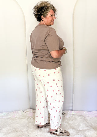A woman stands in profile wearing a brown short-sleeve top and white pants with pink polka dots, standing on a white faux-fur rug in front of a white studio backdrop with arched panels.