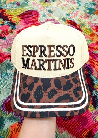 A baseball cap held up by a hand, featuring a cream-colored crown with 'ESPRESSO MARTINIS' embroidered on the front, and a brown leopard-print visor with white piping.