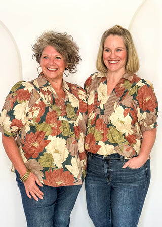 Two women standing side by side wearing matching short-sleeve button-up shirts with a large autumn leaf pattern in rust, beige, and olive green, paired with blue jeans, against a white arched backdrop.