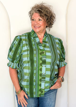A woman with curly gray hair stands against a white arched backdrop, wearing a green and light blue patterned short-sleeve button-up shirt with blue jeans.