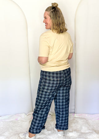 A woman stands with her back to the camera wearing a pale yellow t-shirt and dark blue plaid pajama pants, standing on a white faux fur rug in front of white arch backdrops.