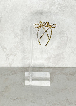 Gold bow-shaped earrings on a clear stand against a marble background