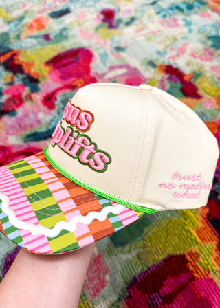 A cream-colored baseball cap with a vibrant multicolor plaid brim and green trim, held by a hand against a colorful floral background. The front of the cap features pink and green text reading 'muscle lifts', and the side has pink script text reading 'trust no matter what'.