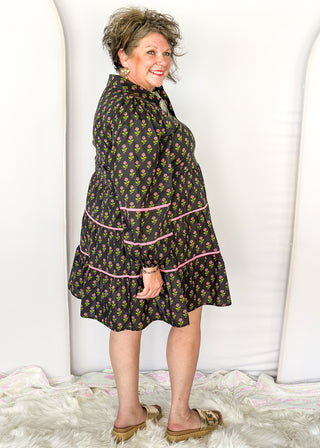 Long sleeve black knee-length dress. Pink and green floral design. Tiered with pink piping detail. Buttons on top.