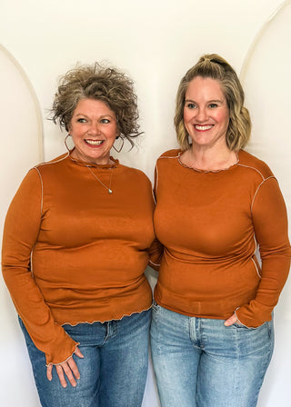 Two women standing side by side wearing matching rust-orange long-sleeve mock neck tops and blue jeans, posing in front of a white arched backdrop.