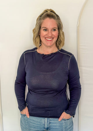 A smiling woman with blonde hair tied back stands against a white arched backdrop, wearing a dark navy long-sleeve top with light piping details and light blue jeans.