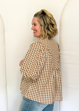 A woman wearing a brown and beige gingham long-sleeve blouse and blue jeans, standing in a white studio setting with arched backdrops.