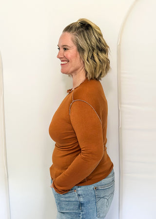 A woman with blonde hair is shown in profile wearing a fitted, long-sleeve rust-orange top and light blue jeans, standing against a white studio backdrop with arch-shaped supports.