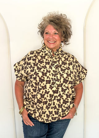 A smiling woman with curly gray hair stands against a white arched wall, wearing a short-sleeve leopard-print blouse with blue jeans.