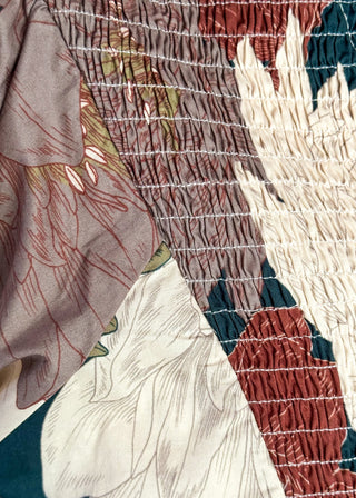 A close-up of a smocked fabric garment or textile panel with a floral print. The fabric features horizontal compression stitching in white over a multicolor pattern of dusty rose, cream, rust red, and dark teal.