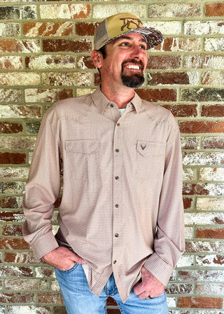 A smiling man wearing a light beige long-sleeve button-up shirt, blue jeans, and a tan baseball cap stands against a rustic brick wall.