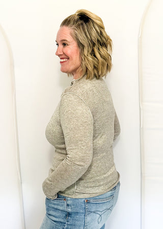 A woman with blonde hair tied back is shown from the side, wearing a light heather gray long-sleeve knit top and blue jeans, standing against a white studio backdrop with arched panels.