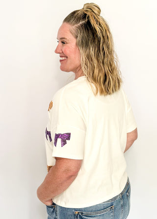 Short sleeve white top featuring scattered sequin bows in gold and purple