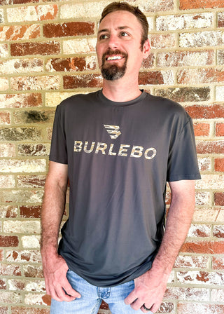 A man wearing a dark gray t-shirt with a distressed gold 'BURLEBO' logo and graphic, standing against a red and beige brick wall.