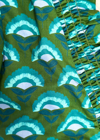 Close-up of a green fabric with a repeating teal and light blue fan-shaped floral pattern, featuring a gathered ruffled trim along one side.