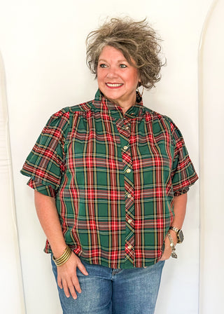 A woman with short, curly gray hair stands against a white studio backdrop wearing a green, red, and black plaid short-sleeve button-up shirt with blue jeans.