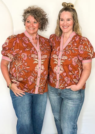 two women standing in a rust, pink and off white button up top and jeans.