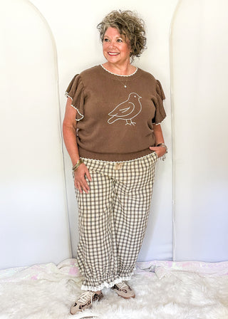A woman modeling a brown short-sleeve knit top with a white bird graphic and beige-and-white gingham pants, standing against a white arched backdrop on a white faux-fur rug.