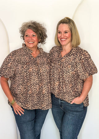 Two women standing side-by-side wearing matching leopard print short-sleeve button-up shirts and blue jeans, posing against a white arched backdrop.