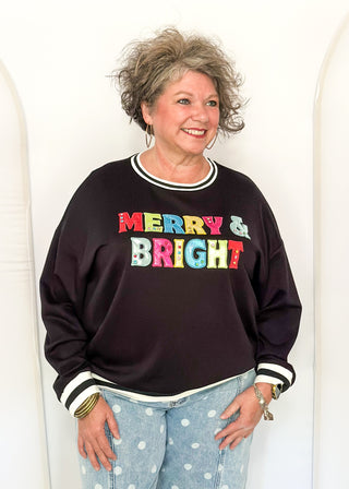 A smiling woman with short gray hair stands against a white arched backdrop, wearing a dark brown long-sleeve sweatshirt with 'MERRY & BRIGHT' printed across the chest in colorful letters, paired with light blue polka-dot jeans.