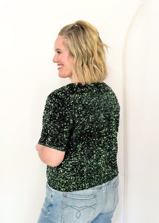 A woman with blonde hair is shown from the back wearing a dark green sequined short-sleeve top and light blue jeans, standing against a plain white studio background.