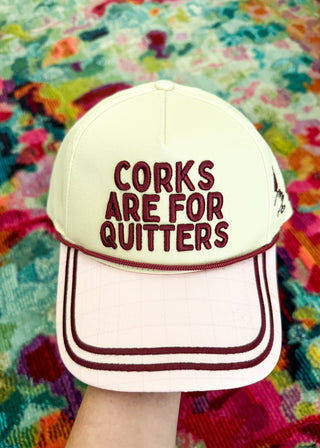 A cream-colored baseball cap with dark red embroidered text reading 'CORKS ARE FOR QUITTERS' on the front, held by a hand against a colorful, patterned background.