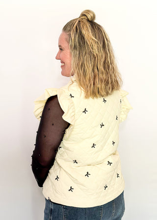 Ivory quilted puffer vest-ruffle sleeve, front pockets, snap closure. Small black bows embroidered throughout.  