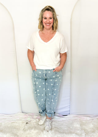 A woman stands indoors wearing a white short-sleeve V-neck T-shirt and light blue jeans with white polka dots. She is standing on a white faux-fur rug in front of a white studio backdrop with arched panels.