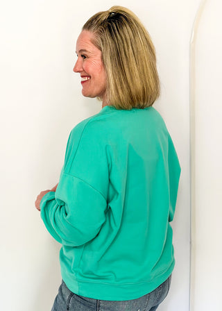 side view of woman wearing a long sleeve top in aqua with watercolor tree graphic and gemstone details.