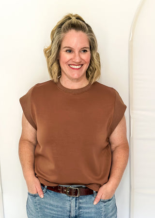A smiling woman with blonde hair tied back stands against a white studio backdrop, wearing a brown short-sleeve crewneck top and light blue jeans.