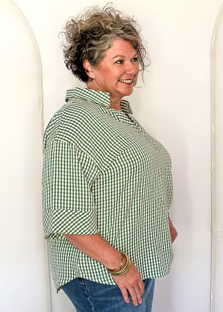 A woman modeling a green and white gingham button-down shirt with three-quarter sleeves, paired with blue jeans, standing against a white studio backdrop with arched panels.
